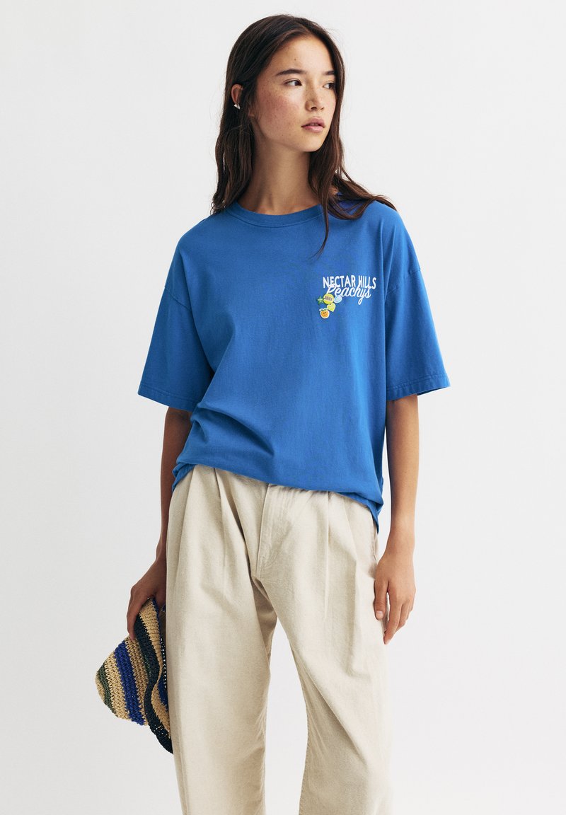 T-shirt en coton bleu avec coupe décontractée, manches courtes, col rond, arborant du texte blanc "NECTAR HILLS" et un petit graphique coloré.