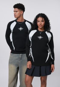Zwarte gebreide truien met witte hoekige accenten en een centraal geborduurd ontwerp. Gecombineerd met lichtdenim broek en een geplooide marineblauwe rok.