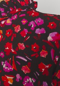 Kwiatowa bluzka na ciemnym tle, z czerwonymi, różowymi i fioletowymi wzorami. Falbaniasty detal przy dekolcie. Miękka tekstura materiału.