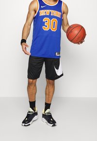 Niebieska koszulka koszykarska z pomarańczowymi napisami i numerem 30. Czarne szorty z białym logo Nike. Czarno-żółte sneakersy, trzymający piłkę do koszykówki.