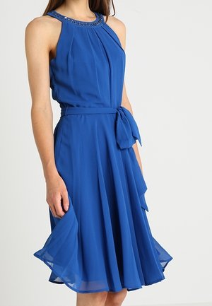 Hihaton kuninkaansininen mekko, jossa helmillä koristeltu kaulus, laskostettu etuosa, vyötärökiristys ja liehuva polvipituinen hame, jota pitää päällään seisova nainen.