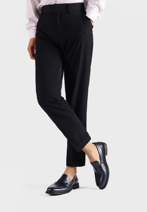 Pantaloni neri con una vestibilità tailored e orlo a sbuffo, abbinati a mocassini blu scuro con una finitura in pelle liscia e dettagli di cucitura discreti.