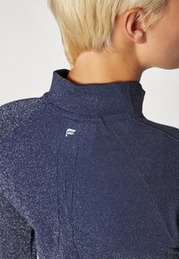 Marineblaues Langarmoberteil mit hohem Kragen, das ein strukturiertes Muster aufweist und ein kleines Logo auf der Rückseite hat, das dezente Glitzereffekte zeigt.