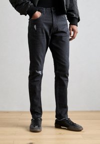 Crne traperice slim fit kroja, s iznosenim zakrpama na oba koljena i pet džepova, u kombinaciji s crnim sportskim tenisicama.