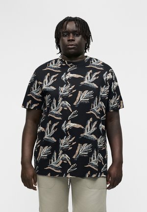 Άνδρας με κοντά dreadlocks φοράει μαύρο T-shirt με μπεζ και λευκά αφηρημένα μοτίβα φύλλων και μπεζ παντελόνι, στέκεται μπροστά σε απλό φόντο.