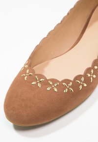 Chaussure plate en daim marron avec un bord festonné et de petits clous floraux dorés le long de l'encolure avant.