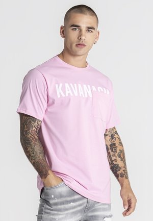 Mladý muž s krátkymi vlasmi a tetovanými rukávmi nosí ružové tričko s nápisom "KAVANAUGH" a svetlosivé opotrebované džínsy, stojí s rukou v kapse.