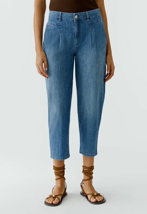 Vrouw die blauw cropped jeans met hoge taille en voorplooien draagt, gecombineerd met bruine platte sandalen met veter, staand tegen een effen achtergrond.