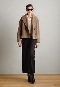 Bruine wolmix dubbellaagse jas, crèmekleurige gebreide top, zwarte wijde broek en glanzende zwarte enkellaarsjes. Matte zwarte zonnebril.