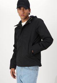 Chaqueta negra impermeable con capucha, cierre frontal de cremallera y dos bolsillos laterales. Lleva puesta sobre una camisa blanca con jeans de color azul claro.