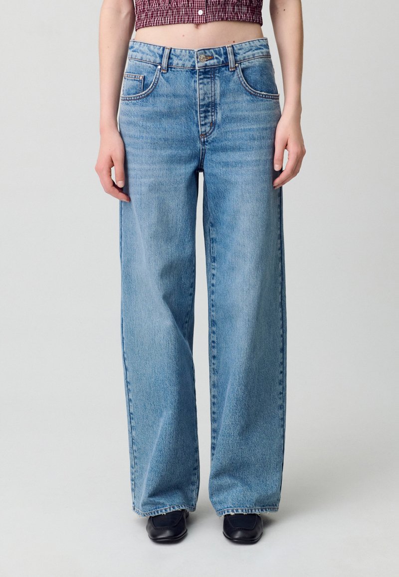 Vaqueros de pierna ancha en azul claro hechos de mezclilla, con cintura alta, cinco bolsillos y cierre de botón. Textura desgastada en los bajos.
