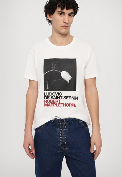 Weißes Baumwoll-T-Shirt mit schwarzem und rotem Text sowie einer schwarz-weißen Blumengrafik. Kombiniert mit dunklen Jeans aus Denim mit Schnürdetails.