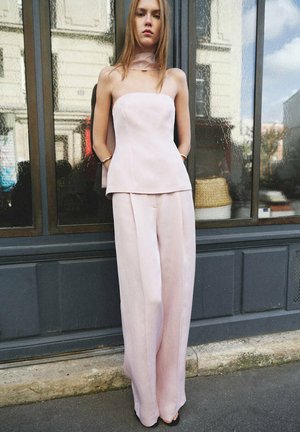 Young woman wearing light pink strapless top with matching wide-leg pants standing against a reflective window outdoors.