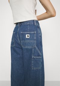 Džínové cargo kalhoty s vysokým pasem v tmavě modré barvě. Obsahují několik kapes, nášivku s logem a volný, pohodlný střih.