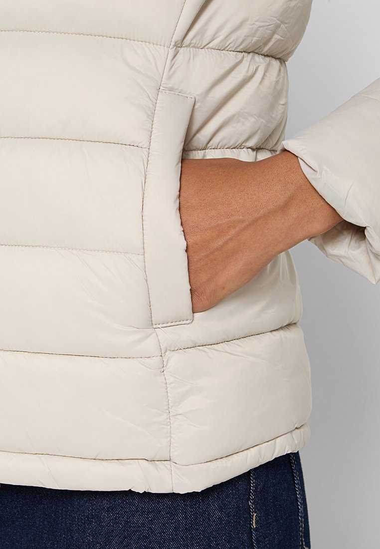 Giacca puffer di un beige chiaro con design a trapuntatura verticale, dotata di una tasca laterale e polsini elasticizzati. Tessuto liscio e resistente all'acqua.