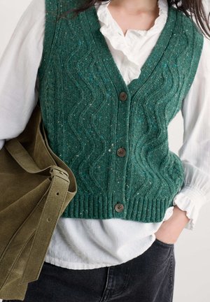 Personne portant un gilet vert en maille torsadée boutonné sur un chemisier blanc à volants, avec un pantalon noir et un sac à bandoulière en daim vert.