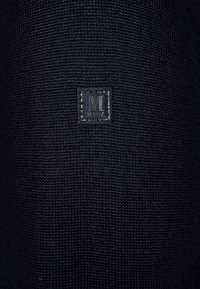 Schwarzer Strickstoff mit einer strukturierten Oberfläche und einem quadratischen Lederpatch, auf dem der Buchstabe 'M' in einer kontrastierenden Farbe gestickt ist.