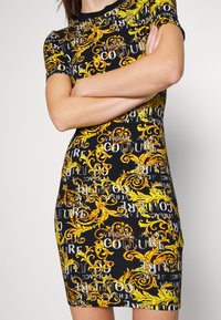 Robe noire ajustée mettant en avant un audacieux motif baroque jaune et blanc avec le texte "VERSACE COUTURE" dans diverses orientations.