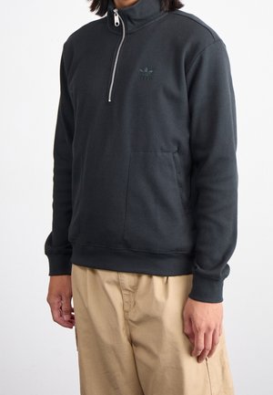 Dunkelgrauer Fleece-Pullover mit hohem Kragen, halblangem Reißverschluss, gerippter Textur und Logo auf der Brust, kombiniert mit beigen Hosen.