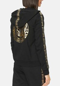 Zwarte hoodie met gouden accenten, voorzien van een grafisch skeletontwerp op de rug en tekst langs de mouwen. Zacht, gestructureerd stof.