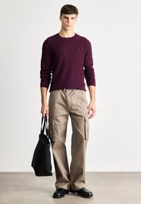 Bordeauxroter Strickpullover und beige Cargo-Hosen, gehalten mit einer schwarzen Tragetasche. Die Hose verfügt über Seitentaschen, und die Schuhe sind schwarz mit einer glatten Oberfläche.