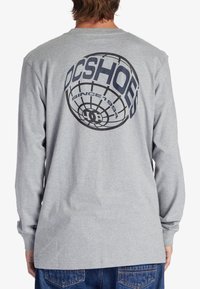 Long-sleeve gray t-shirt featuring a large black graphic print of a globe with "DC SHOES" and "SINCE 1994" text on the back.