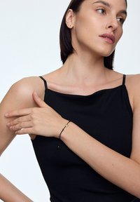 Czarny prążkowany top z cienkimi ramiączkami. Modelka nosi czarną bransoletkę z cordu z złotą pętlą i małym złotym akcentem.