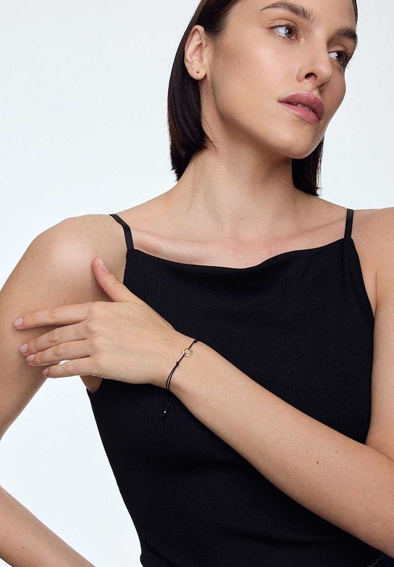 Czarny prążkowany top z cienkimi ramiączkami. Modelka nosi czarną bransoletkę z cordu z złotą pętlą i małym złotym akcentem.