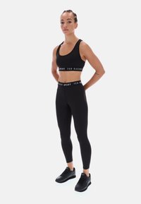 Juodas sportinis komplektas, kuriame yra trumpas viršus su "TED BAKER SPORT" logotipu ir aukštu liemeniu leggingai, derinami su juodais sportiniais batais.