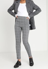 Nainen, jolla on yllään harmaat ruudulliset slim-fit-housut, valkoinen paita, harmaa bleiseri ja mustat nahkakengät, seisoo kädet lanteilla.