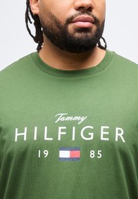 Άνδρας που φοράει πράσινο μπλουζάκι Tommy Hilfiger με λογότυπο, σημαία και το κείμενο "1985" στο στήθος, αποκομμένο στον λαιμό και στους ώμους.