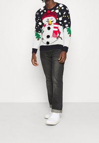 Strickpullover mit schwarzem Schneemann und weißen, roten und grünen Akzenten; mit Pompons; kombiniert mit dunklen Jeans und weißen Sneakers.
