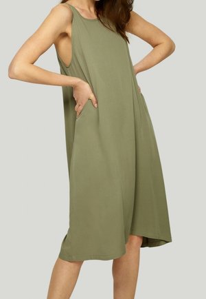 Kobieta ubrana w luźną, bezrękawnikową sukienkę oliwkową z głębokimi bocznymi kieszeniami, stoi z rękami w kieszeniach na jednolitym tle.