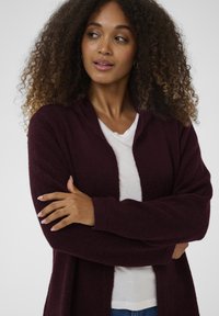 Bordowy kardigan z otwartym przodem wykonany z miękkiego, teksturowanego materiału, z długimi rękawami i luźnym krojem, noszony na białej koszulce.