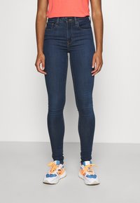 Pantalones vaqueros ajustados de denim azul con una cintura alta, costuras mínimas y una textura suave, combinados con zapatillas deportivas blancas y azules con acentos naranjas.