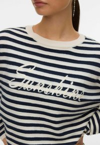 Femme portant un pull rayé bleu marine et blanc avec le mot « Sincerely » brodé sur la poitrine, et une délicate boucle d'oreille pendante visible.