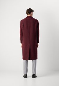 Palton din lână bordeaux cu o siluetă ajustată, ventral spate și umeri structurați, purtat cu pantaloni gri deschis și pantofi negri.