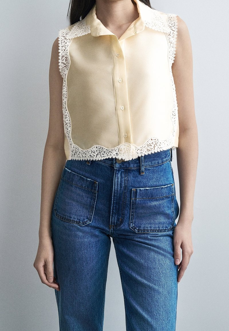 Femme portant un chemisier sans manches jaune pâle à boutons avec bordure en dentelle et un jean bleu taille haute avec poches plaquées devant.