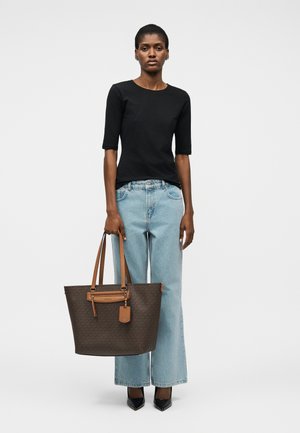 Ženska, oblečena v črno majico s kratkimi rokavi, svetlo modre široke kavbojke, črne koničaste čevlje, drži veliko rjavo usnjeno torbo z rjavimi ročaji.