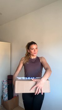 Kartonnen doos in beide handen gehouden, bruin van kleur, met een zichtbaar wit verzendetiket. Persoon draagt een nauwsluitend bruin shirt en zwarte legging.