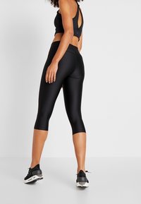 Czarny zestaw sportowy składający się z crop topu z okrągłym wycięciem z tyłu oraz legginsów do połowy łydki o gładkiej, błyszczącej fakturze. Czarne sneakersy.