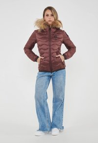 Chaqueta acolchada marrón con capucha forrada de piel, cierre de cremallera y bolsillos laterales. Combinada con vaqueros azules desgastados y zapatos blancos.