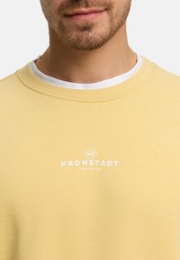 Lys gul bomullsgenser med rund hals, med hvit logo og tekst i midten. Myk tekstur og avslappet design.