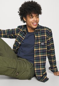 Camisa de flanela com padrão de xadrez em azul, verde e amarelo; dois bolsos no peito. T-shirt azul-marinho por baixo; calças de carga verde-oliva com detalhes de costura.