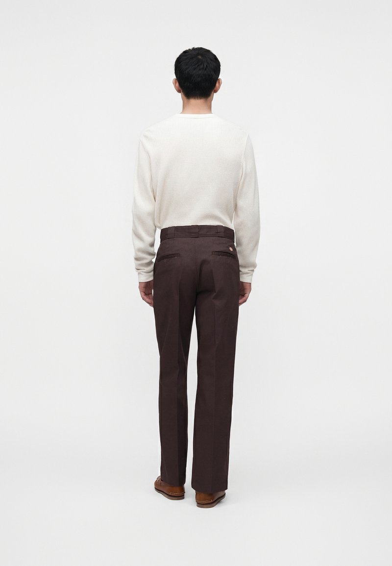Uomo in piedi di spalle, indossa un maglione a coste color crema, pantaloni marrone scuro e scarpe marroni su uno sfondo bianco semplice.