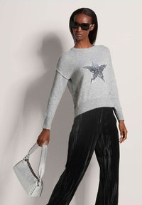 Grauer Pullover mit einem silbernen Paillettenstern-Design, kombiniert mit schwarzen, gefalteten Hosen und einer metallisch-silbernen Handtasche. Model trägt dunkle Sonnenbrillen.