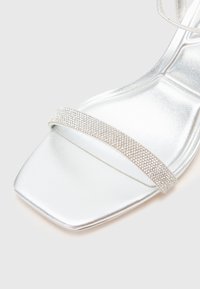 Silver högklackad sandal med fyrkantig tå, med en rem prydd med rhinestones och ett glänsande, slätt syntetmaterial.