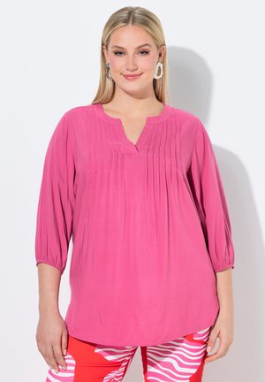 Frau trägt eine hellrosa plissierte Bluse mit Dreiviertelärmeln und rot-weißen gemusterten Hosen, lächelnd mit glattem blondem Haar.