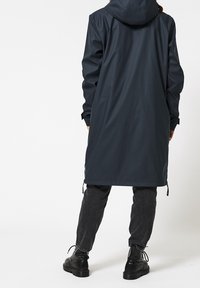 Lång mörkblå vattentät kappa med huva, justerbara dragsnören vid fållen och långa ärmar, kombinerad med svarta byxor och skor.