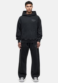 Felpa oversize nera con cappuccio e tasca frontale, abbinata a pantaloni felpati neri dal taglio ampio. Il materiale sembra morbido, con dettagli testuali minimi.